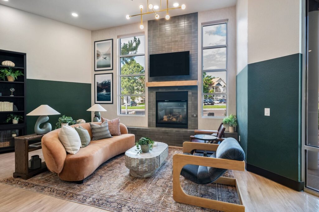 Modern living room with a tan curved sofa, wooden armchair, oval stone coffee table, and a fireplace with a TV above. Large windows and framed art let in natural light. Plants and a rug add cozy touches.