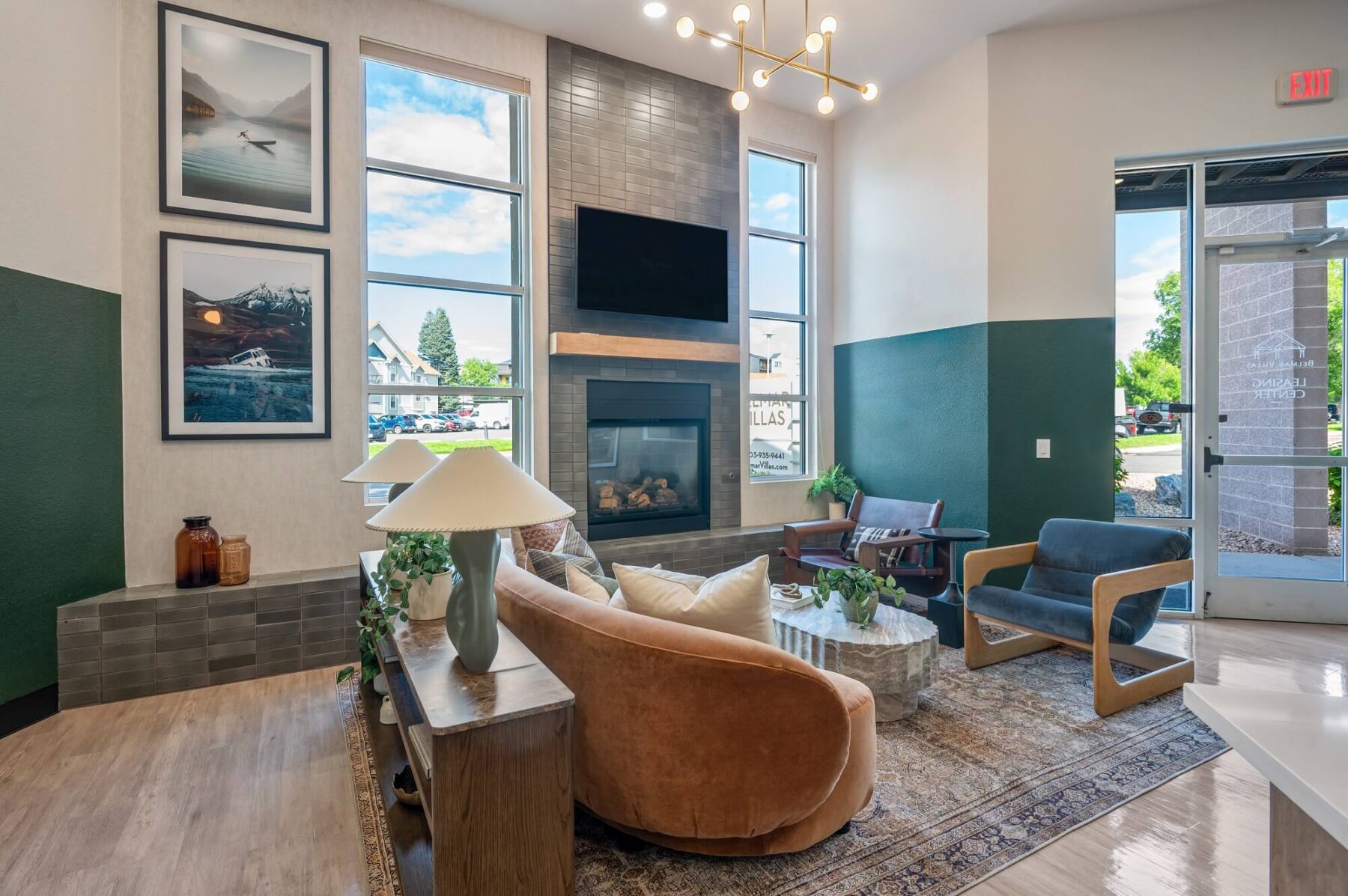 Modern lobby of apartments in Vancouver WA with a tan couch, blue chairs, marble coffee table, fireplace, TV, large windows, framed art, green accents, and potted plants. Natural light fills the stylish and inviting space.